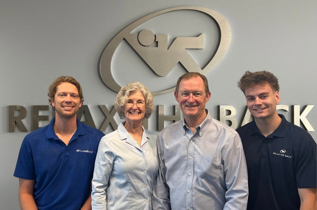 Four people standing together in front of a gray wall with a large metallic logo and the words 'RELAX THE BACK' at Relax the Back.