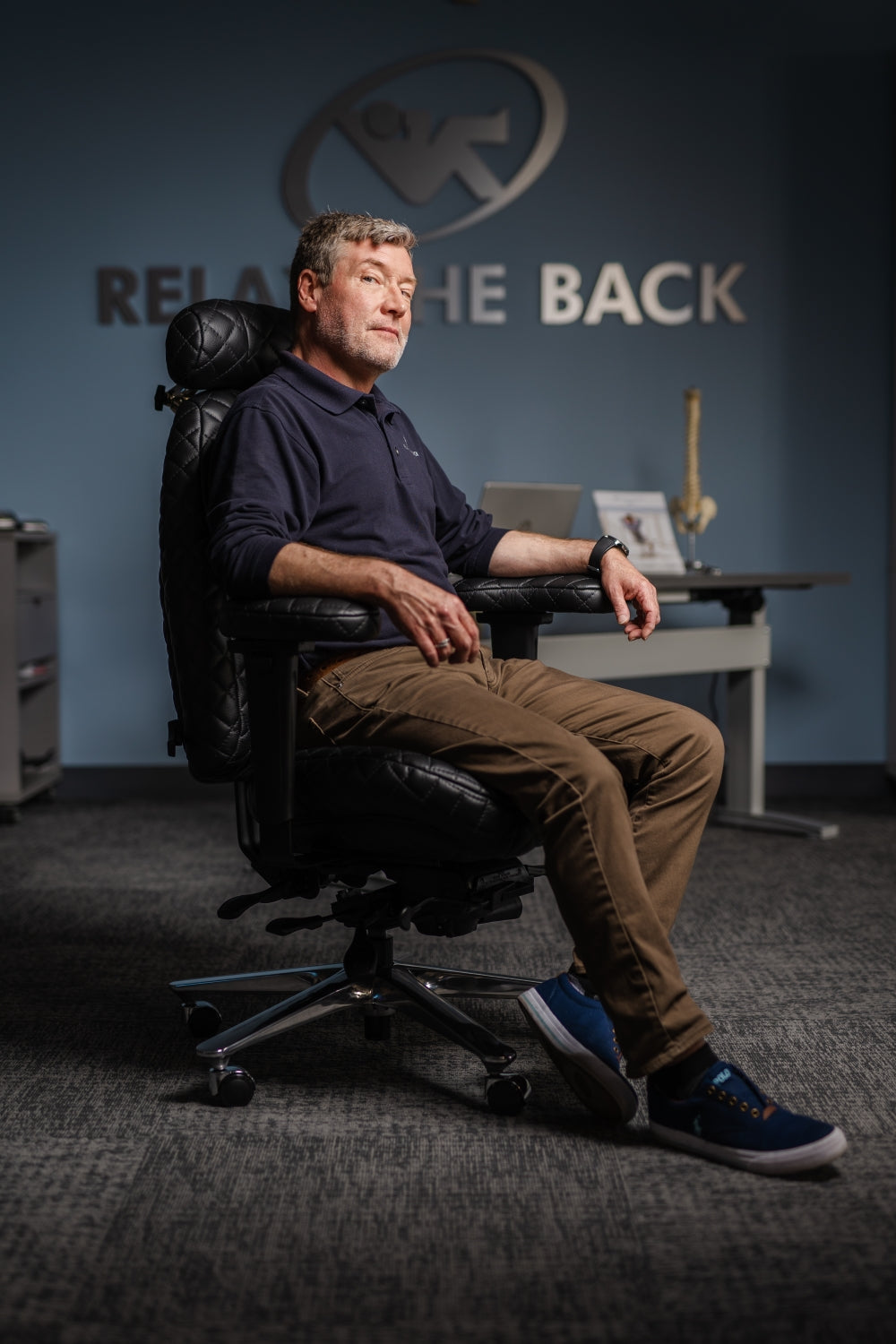 Man sitting in a chair with a blurred background featuring a logo and text.