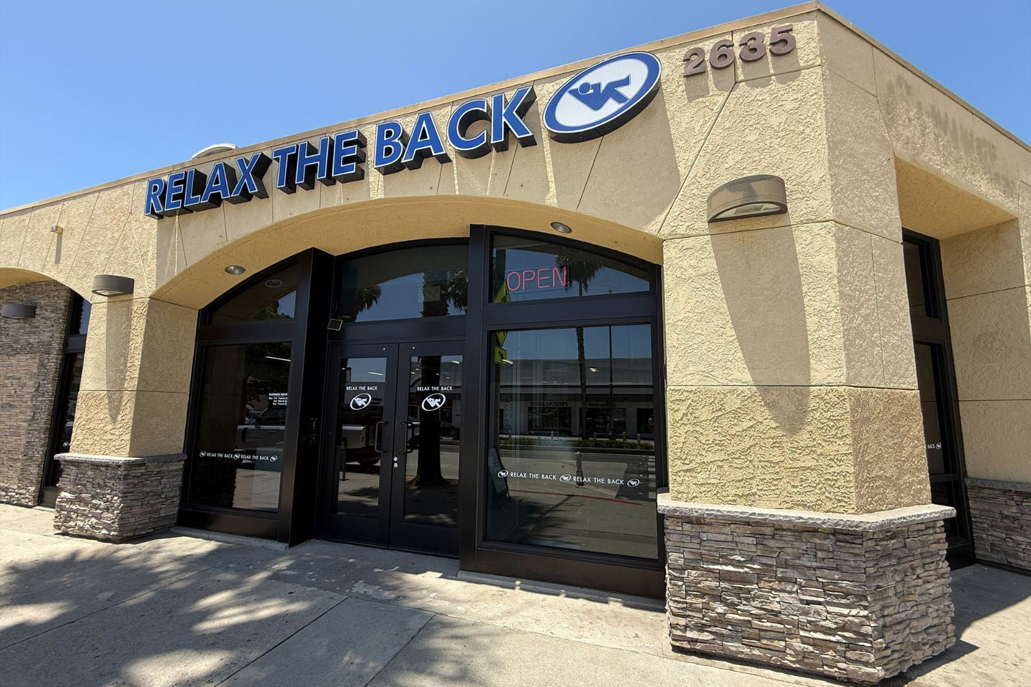 Relax The Back massage therapy clinic exterior, featuring a beige stucco building with a large blue and white logo, glass double doors, and stone accents at the base.