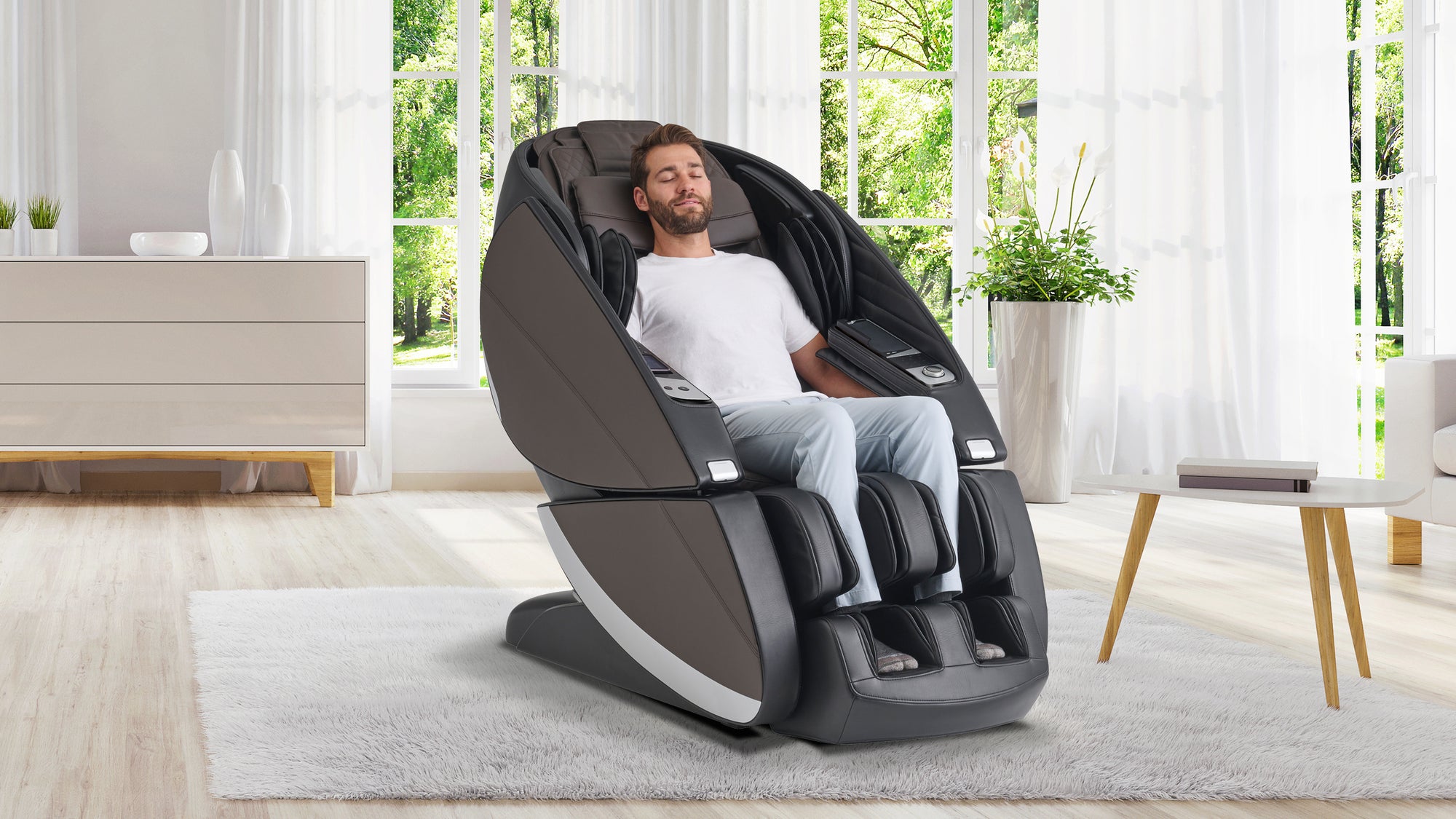 Man relaxing in a modern black massage chair with dark gray side panels, positioned in a bright living room with large windows, white curtains, and natural light. He has his eyes closed, appearing comfortable and at ease, with his legs in the chair’s footrest. A dresser, potted plant, and small table with books are visible in the background.