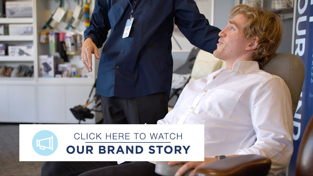 A salesperson wearing a company logo long sleeve shirt helps a man test a recliner in a showroom. The salesperson is demonstrating the reclining features while the man sits in the chair.
