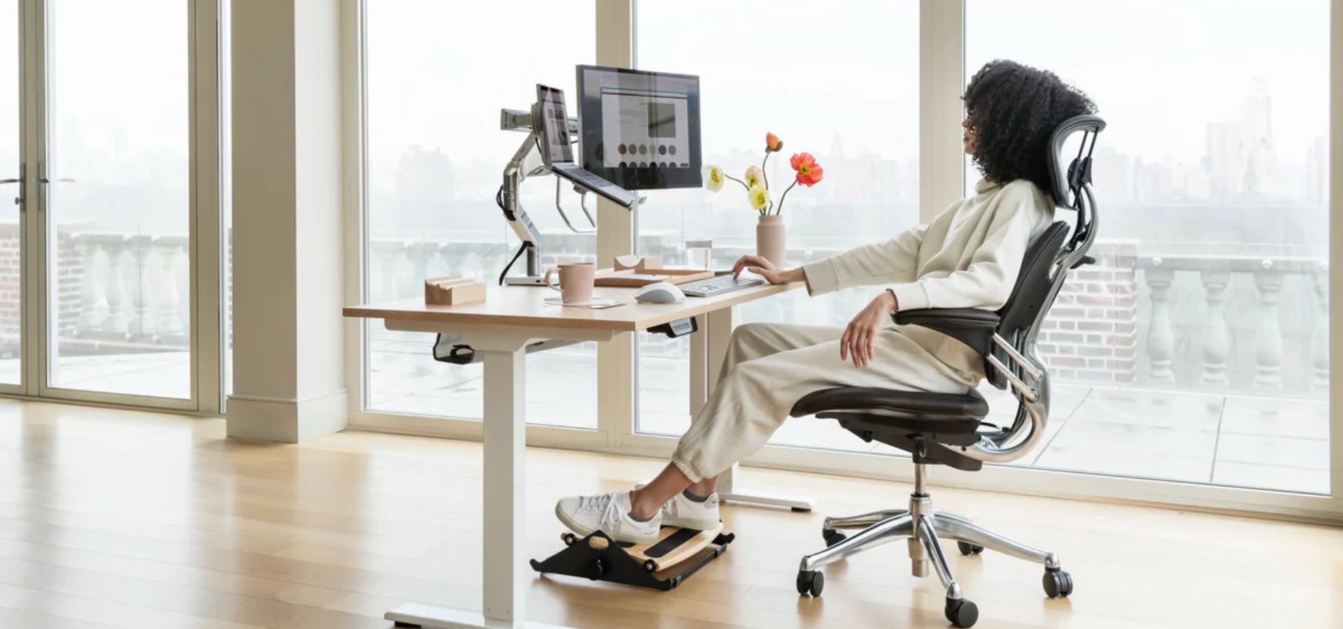 Woman seated in an ergonomic office chair at a standing desk with dual monitors in a bright modern workspace.