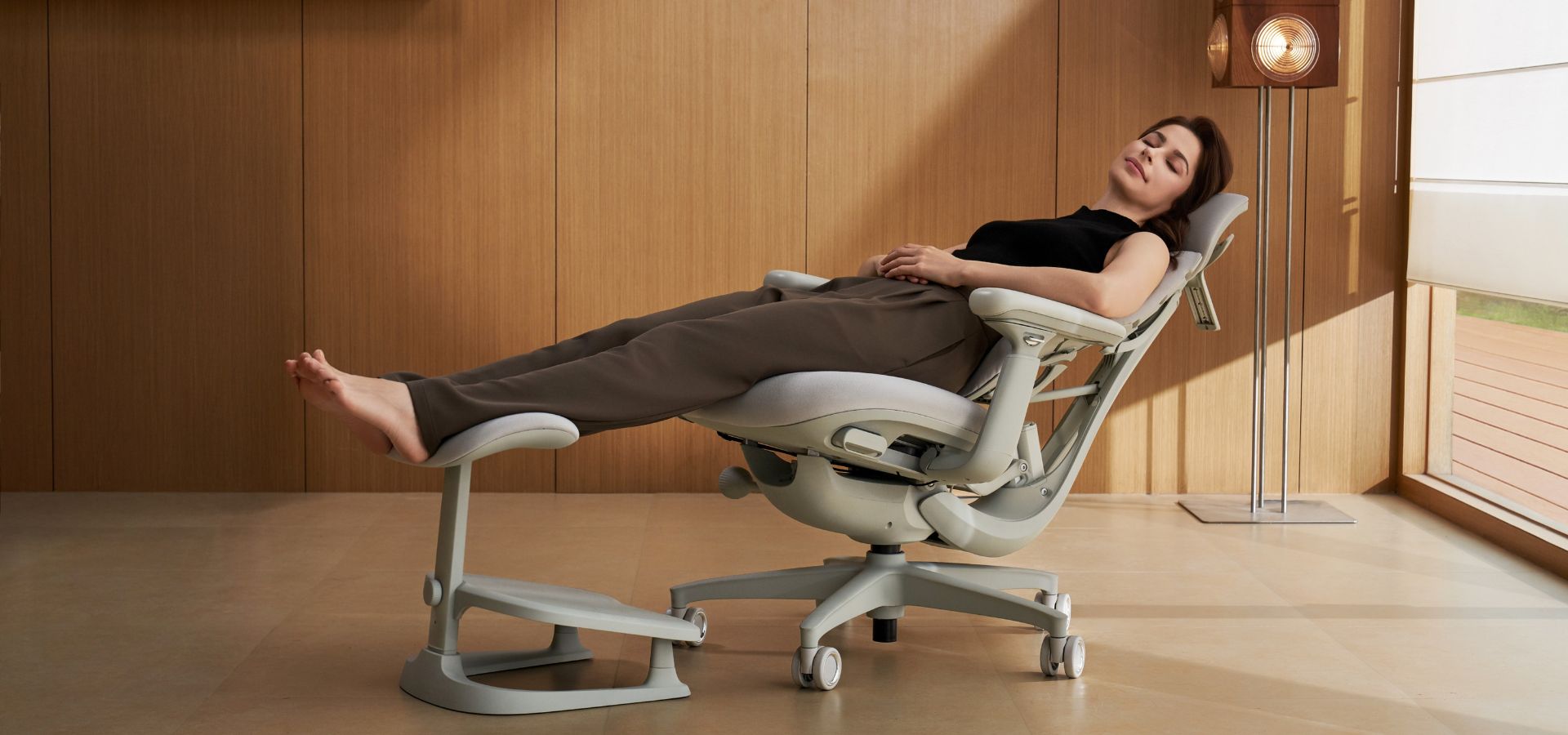 A woman reclines comfortably in an ergonomic office chair with an elevated footrest, resting with her eyes closed in a modern room with warm wood-paneled walls and natural sunlight.