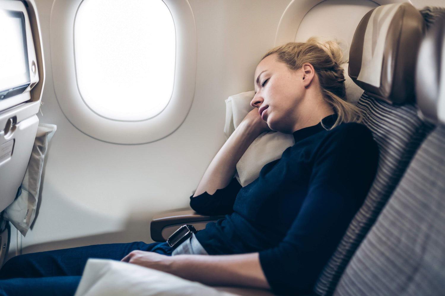 A woman sleeps in an airplane window seat, leaning against a travel pillow during a flight. Sunlight streams through the window beside her, and her seatbelt is fastened as she rests in the cabin.