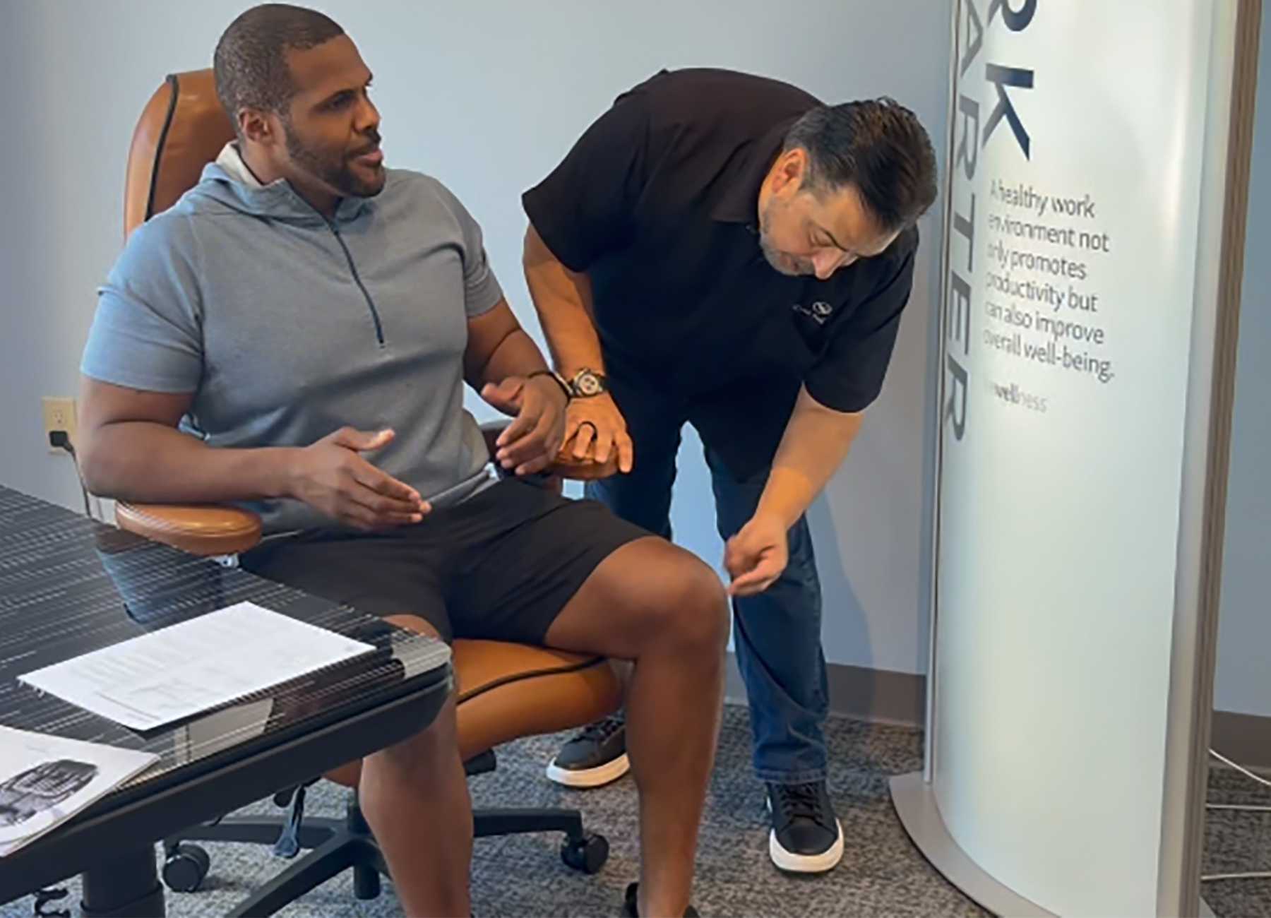 A man in a gray hoodie and black shorts sits in an office chair while another man in a black polo shirt examines his leg. The scene takes place in a modern office environment with a desk, office chair, and a white cylindrical display stand.
