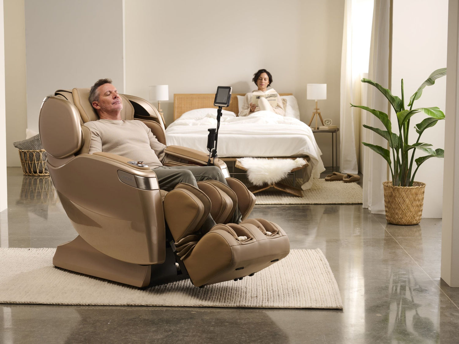 A man using a Cozzia massage chair in a bedroom setting with a woman laying on a bed reading in the background.