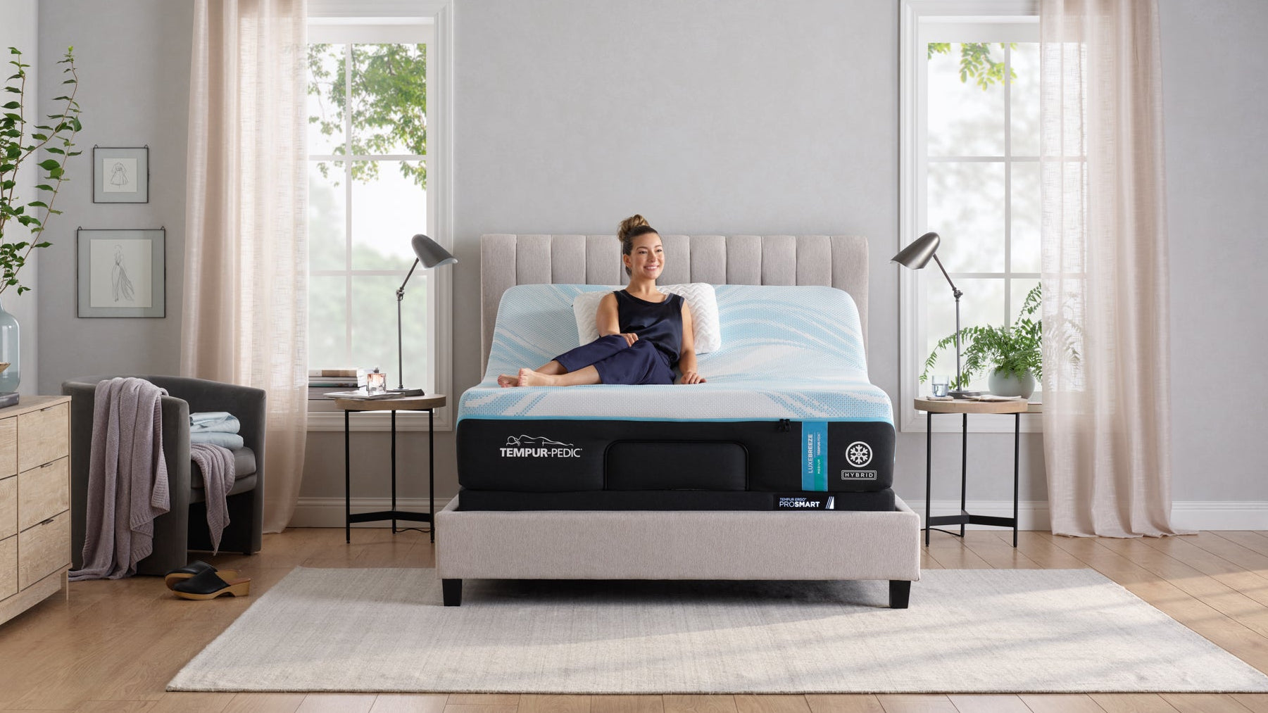 Woman sitting on a bed with a mattress brand logo in a bedroom setting.