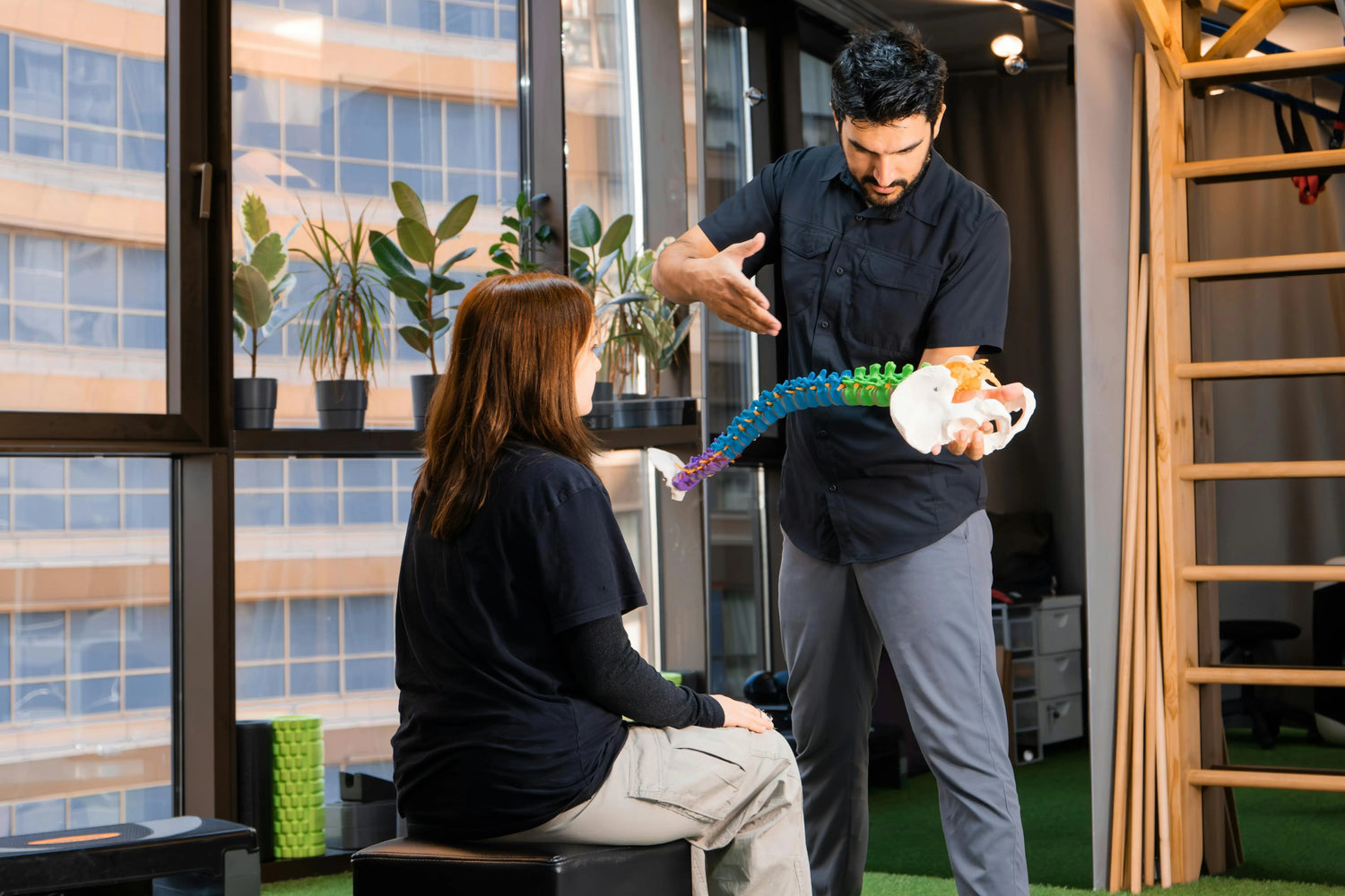 A male chiropractor stands and explains spinal alignment using a colorful 3D model of the human spine and pelvis to a seated female patient.