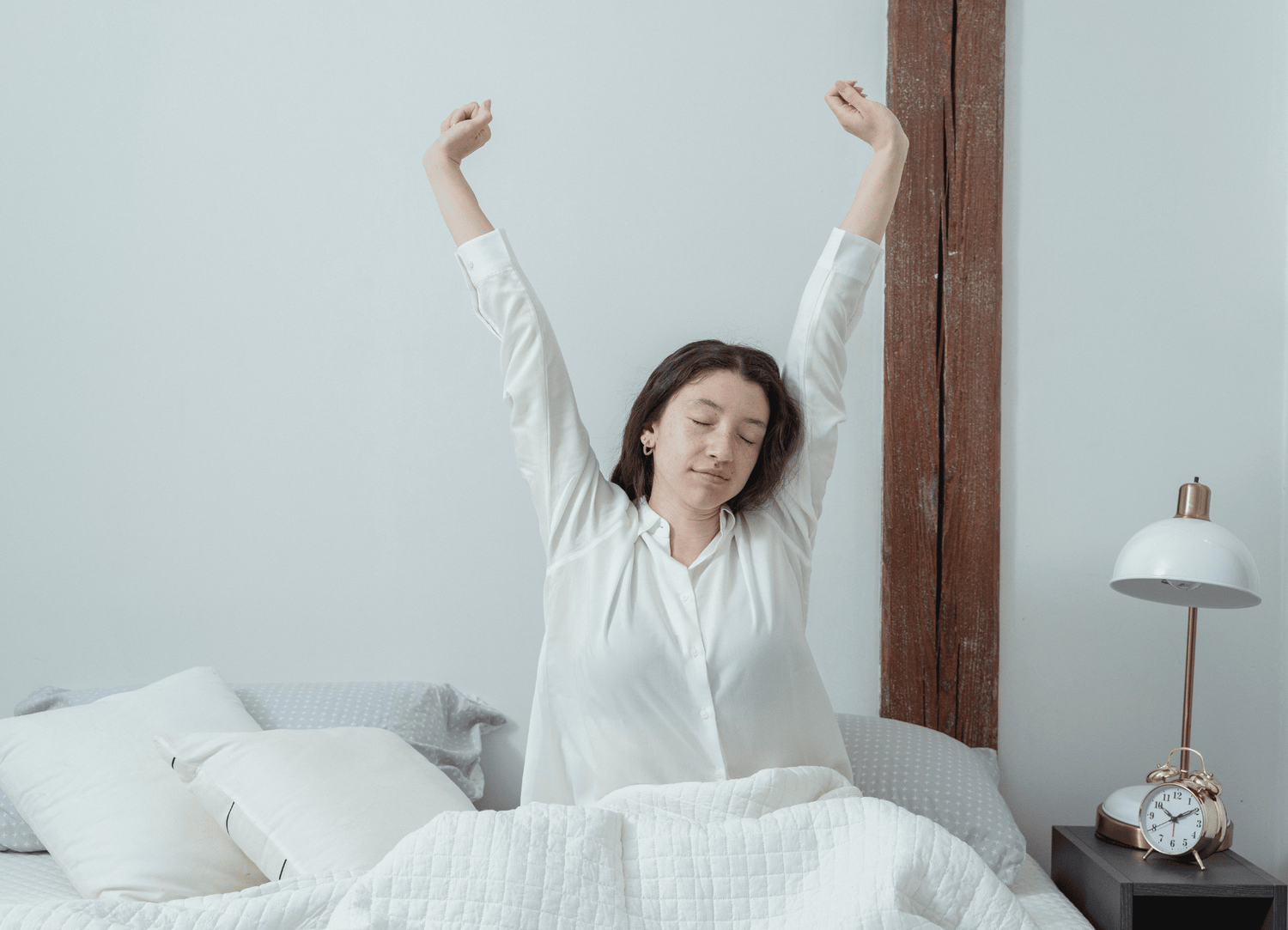 Woman stretching in bed in a bedroom with a clock and lamp on a nightstand.