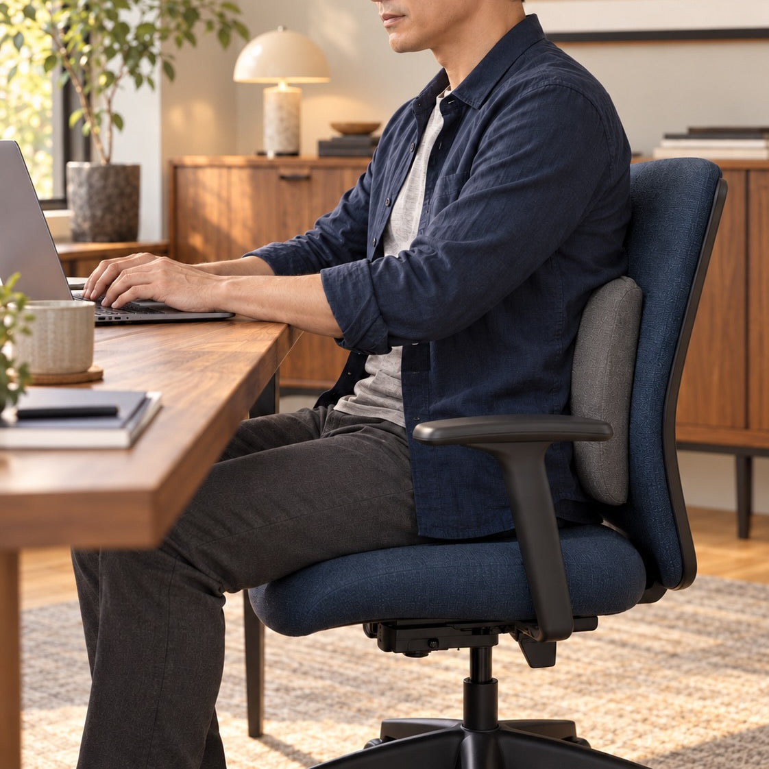 A man sits in a blue upholstered office chair at a wooden desk, working on a laptop in a bright, modern home office.