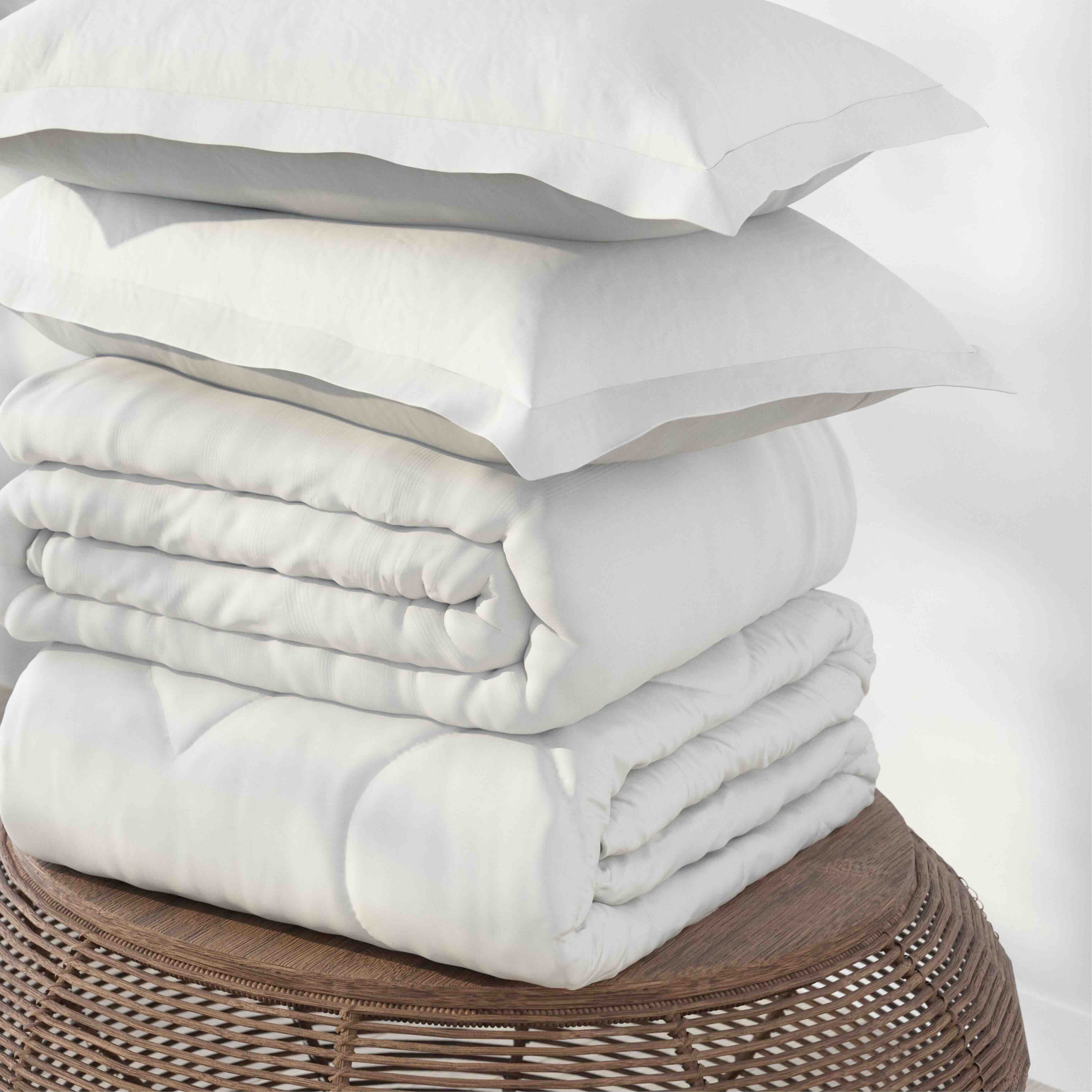 A stack of white bedding, including two pillowcases and duvet cover, displayed on a round woven wicker stool against a plain white background.