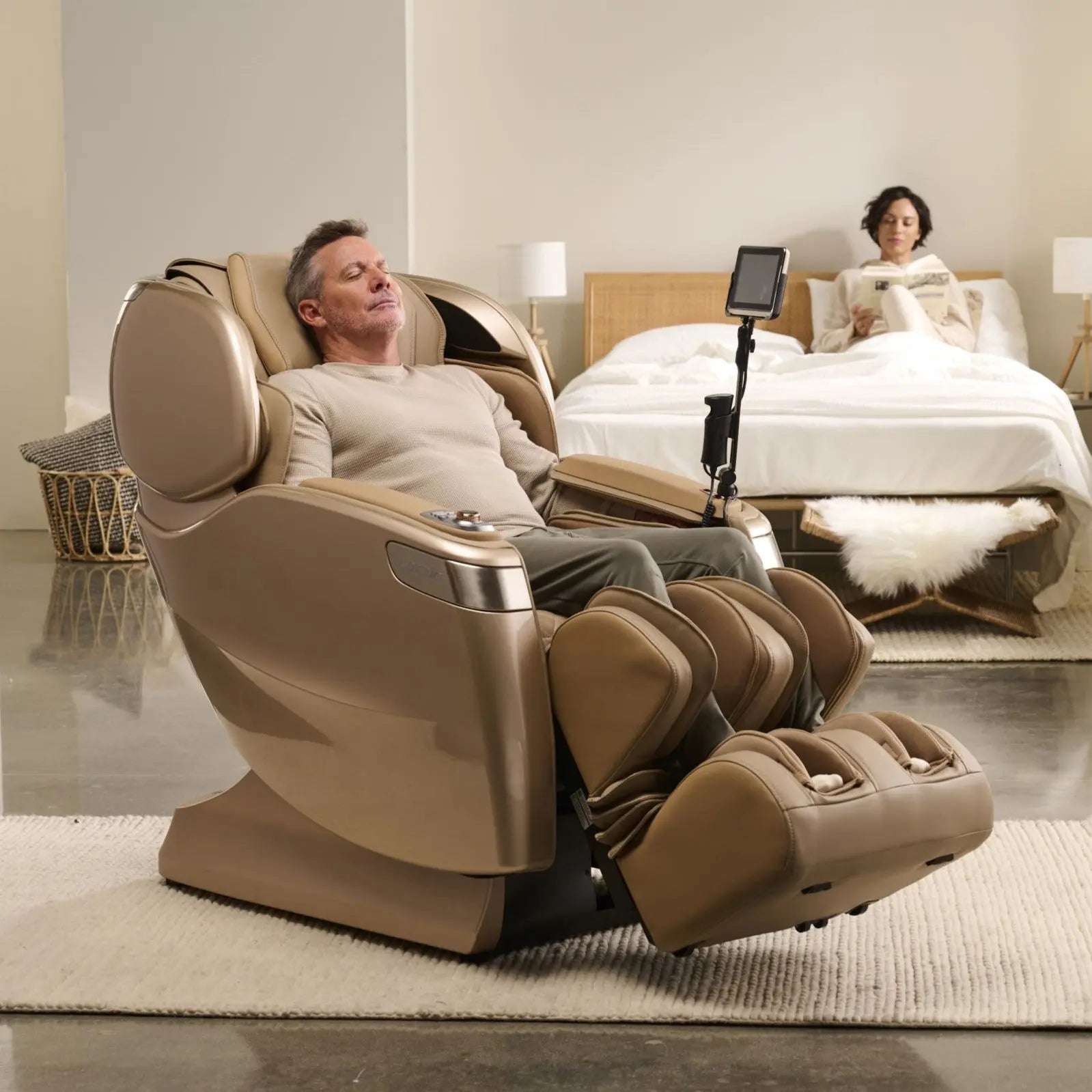 A woman standing beside a man seated in a beige and grey Human Touch Novo Flex massage chair, adjusting the chair settings using the remote.