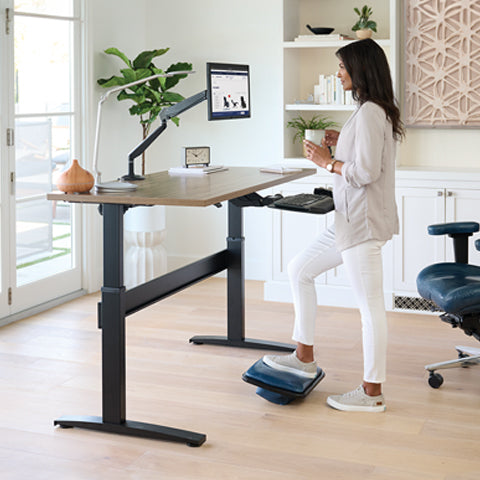 A woman using the Transcend Standing Desk by Relax The Back and a Lifeform padded footrest.