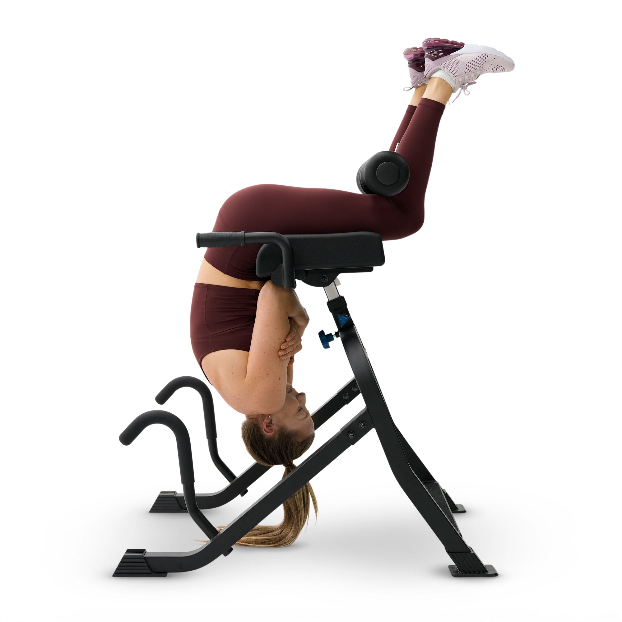 A woman in burgundy athletic wear performs a headstand on a black leg extension gym machine against a clean white background.