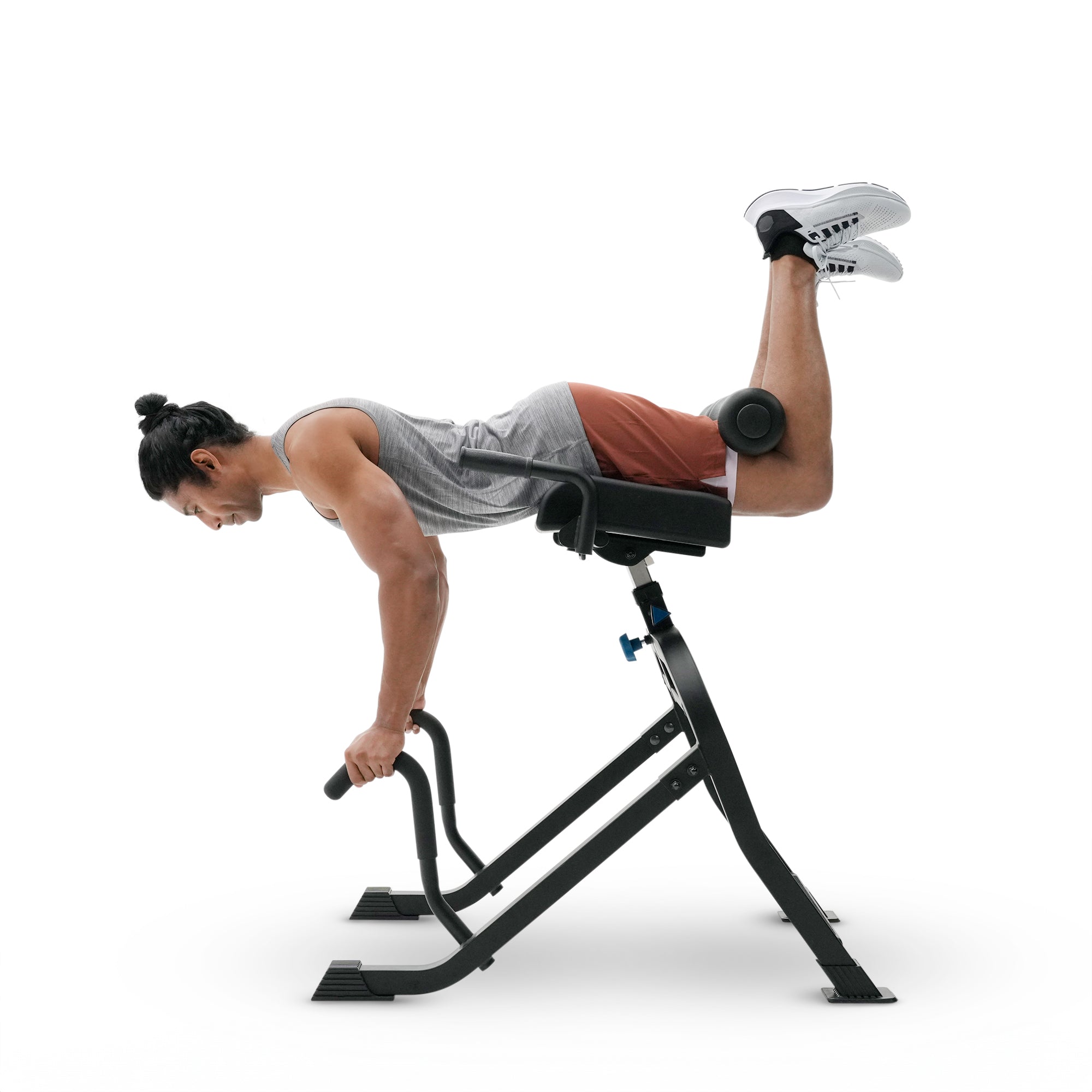 A man performing a hyperextension exercise on a black adjustable leg press/rower gym machine, wearing a gray tank top, rust-orange shorts, and white sneakers, set against a plain white studio background.