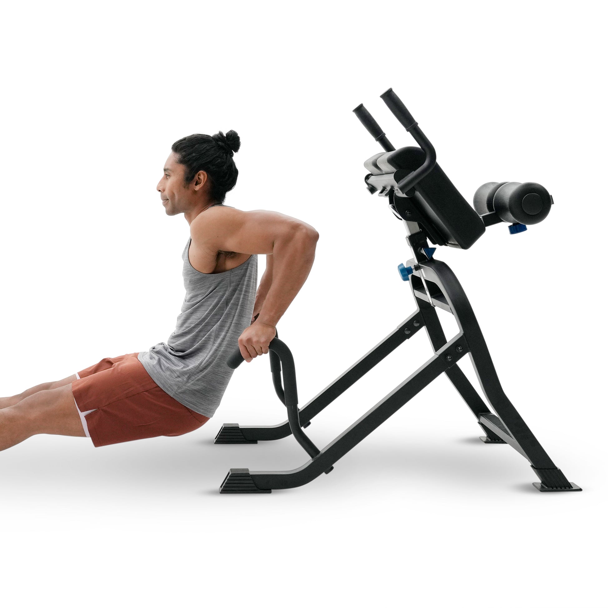 A man in a gray tank top and rust-colored shorts performs a back extension exercise using a black adjustable weight bench with a padded backrest, isolated on a white background.