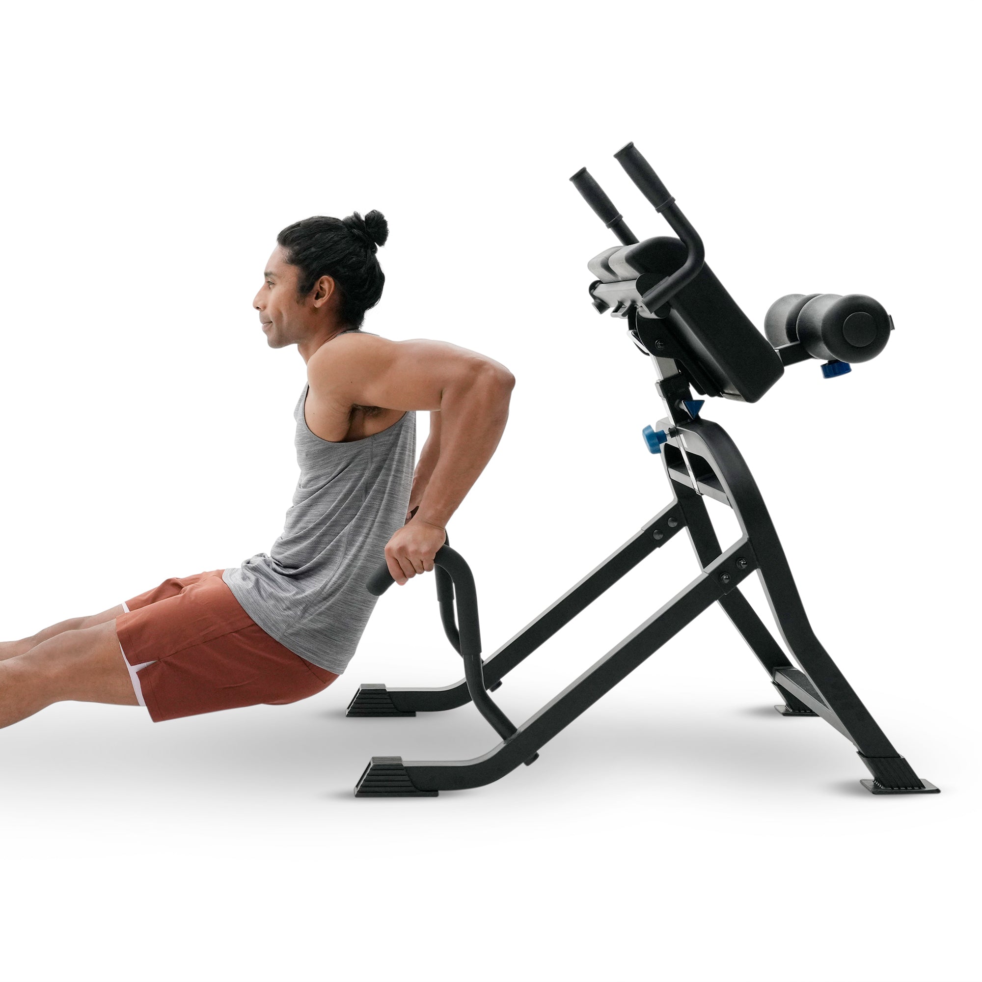 A man in a gray tank top and rust-colored shorts performs a back extension exercise using a black adjustable weight bench with a padded backrest, isolated on a white background.