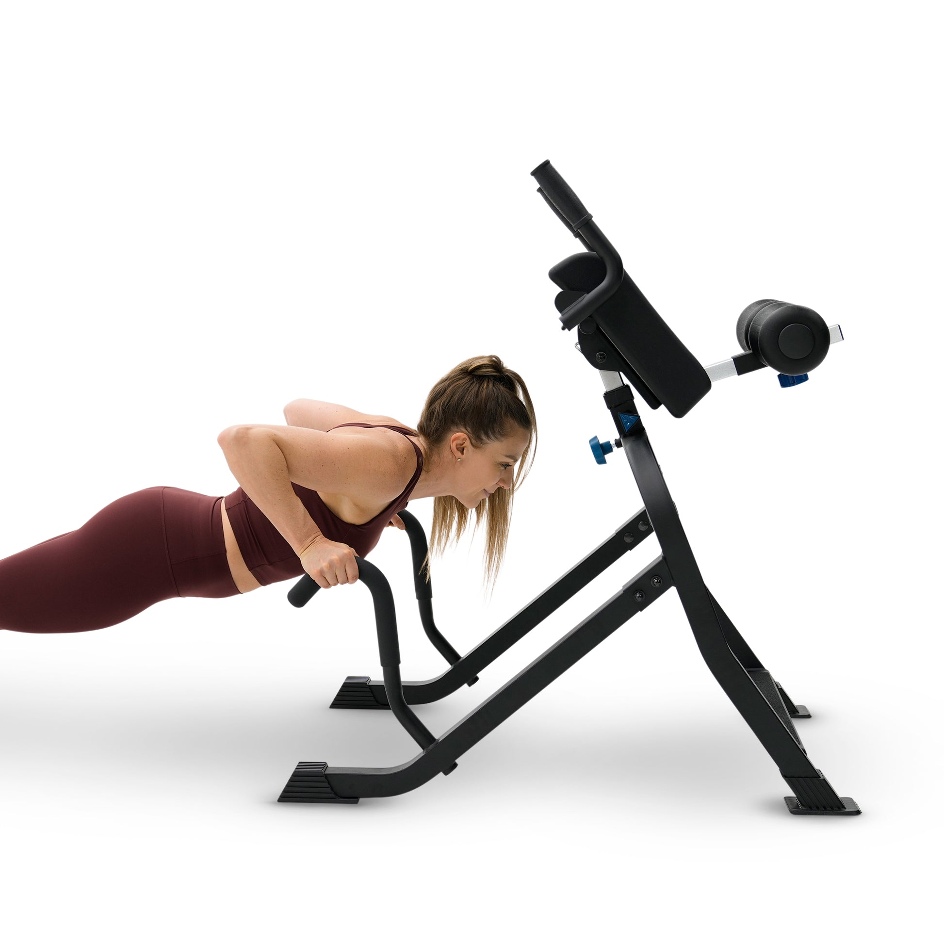 A woman in a maroon sports bra and leggings performs a hyperextension push-up on a black adjustable weight bench with a padded backrest and armrests, isolated on a white background.
