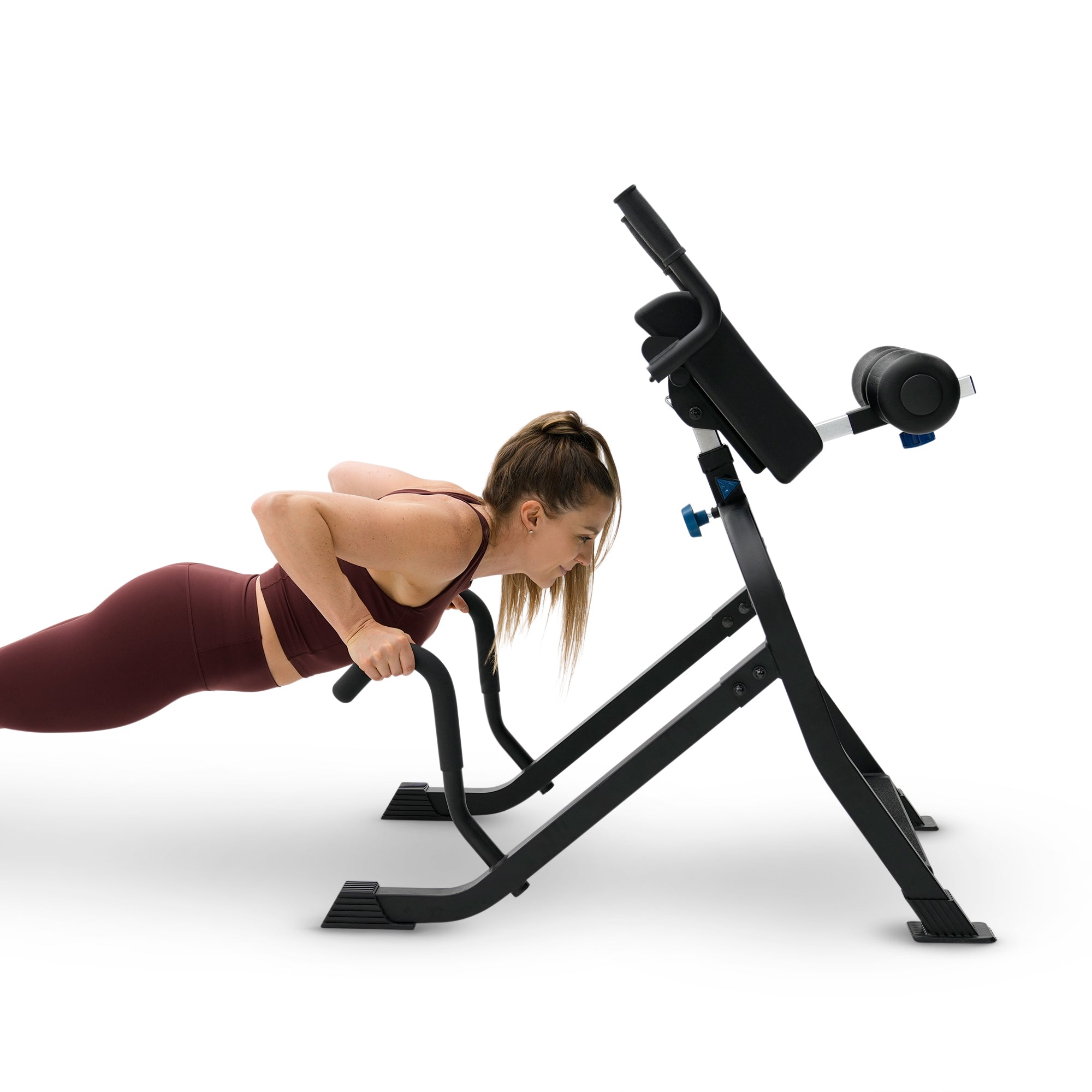 A woman in a maroon sports bra and leggings performs a hyperextension push-up on a black adjustable weight bench with a padded backrest and armrests, isolated on a white background.