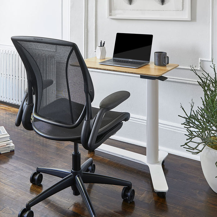 Float Mini Desk in an office setting with an art frame on a wall, a plant to its side and books on the floor.