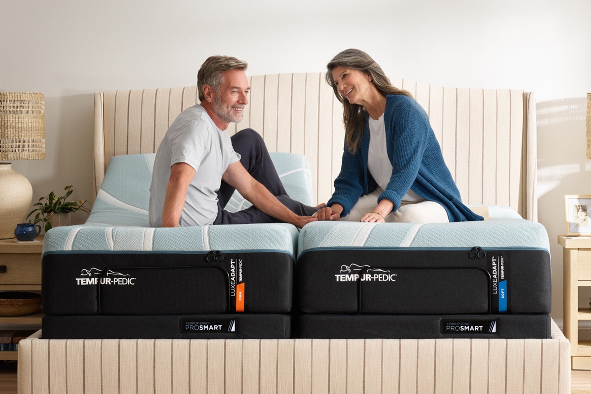A smiling couple sits on split king Tempur-Pedic LuxeAdapt mattresses with ProSmart adjustable bases, holding hands and facing each other in a cozy bedroom setting