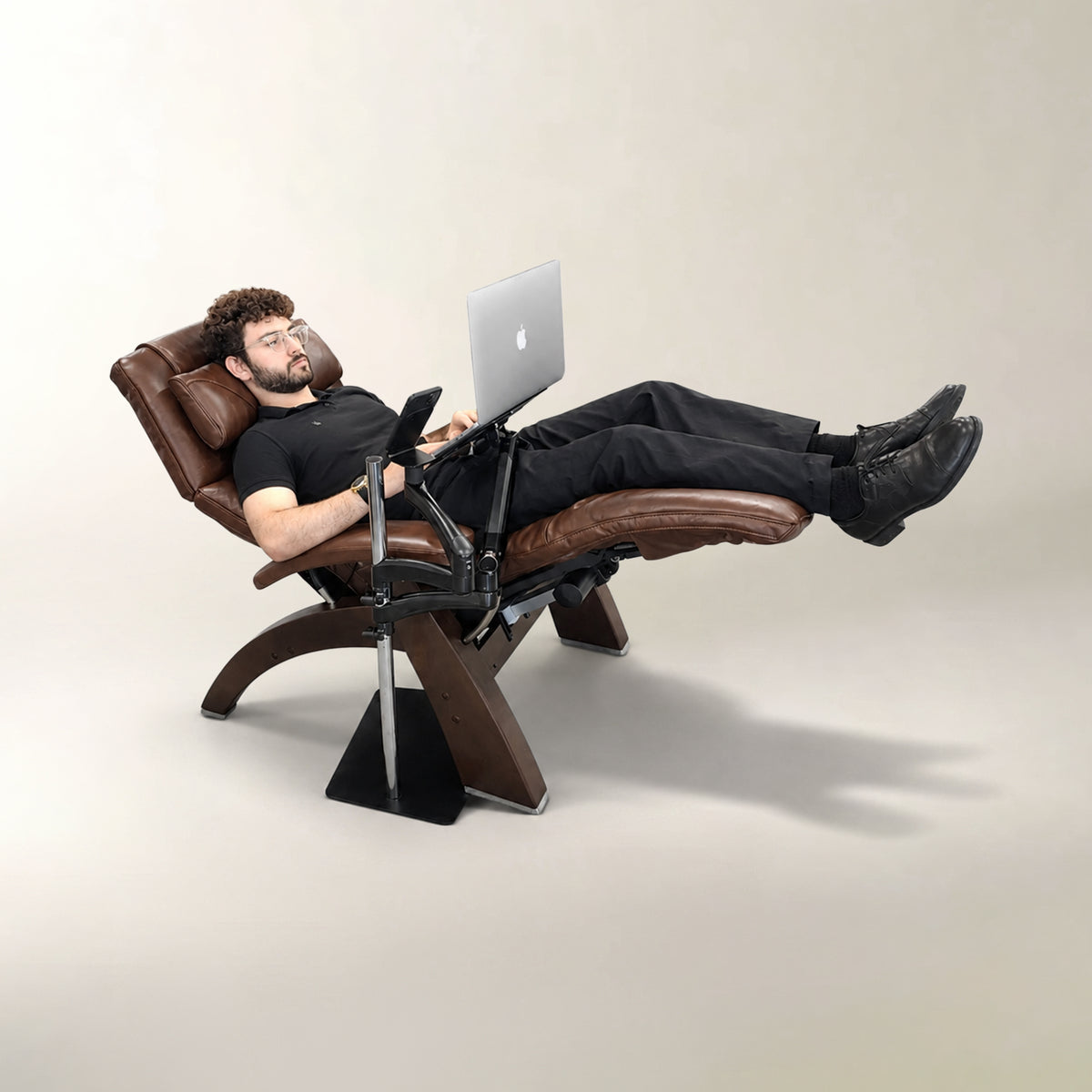 A man reclining in a brown leather ergonomic office chair with a built-in desk and laptop holder, using a silver laptop and holding a smartphone. The chair has a dark wood-look frame and black base, set against a plain beige studio background.