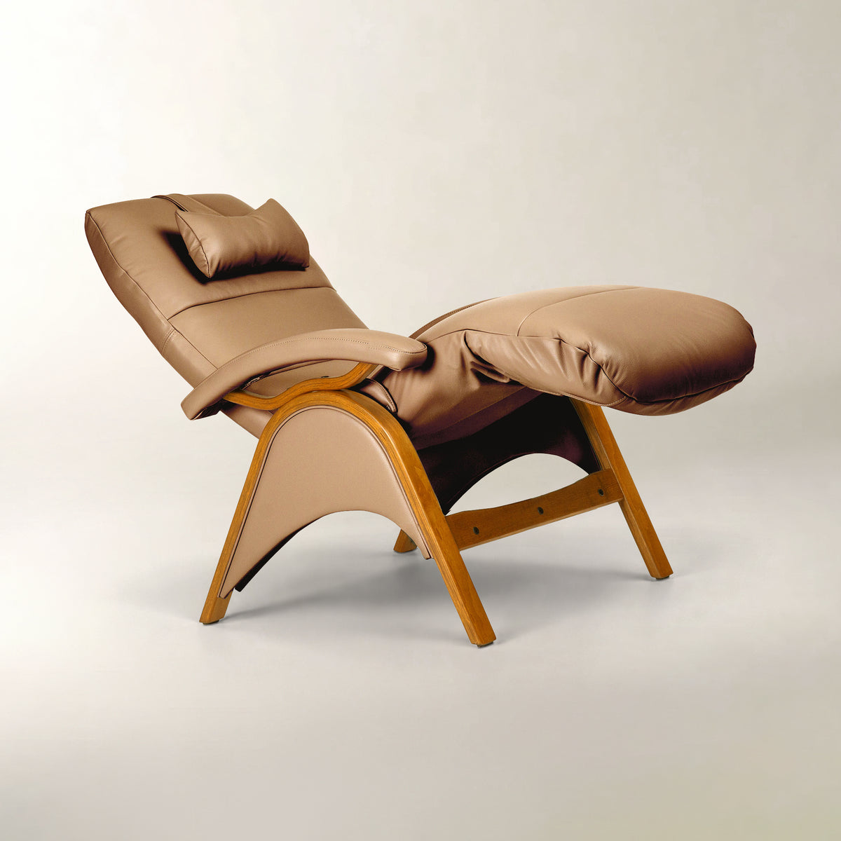A modern tan leather recliner chair with a matching headrest pillow and ottoman, supported by a curved light wood frame, photographed on a neutral light gray studio background.