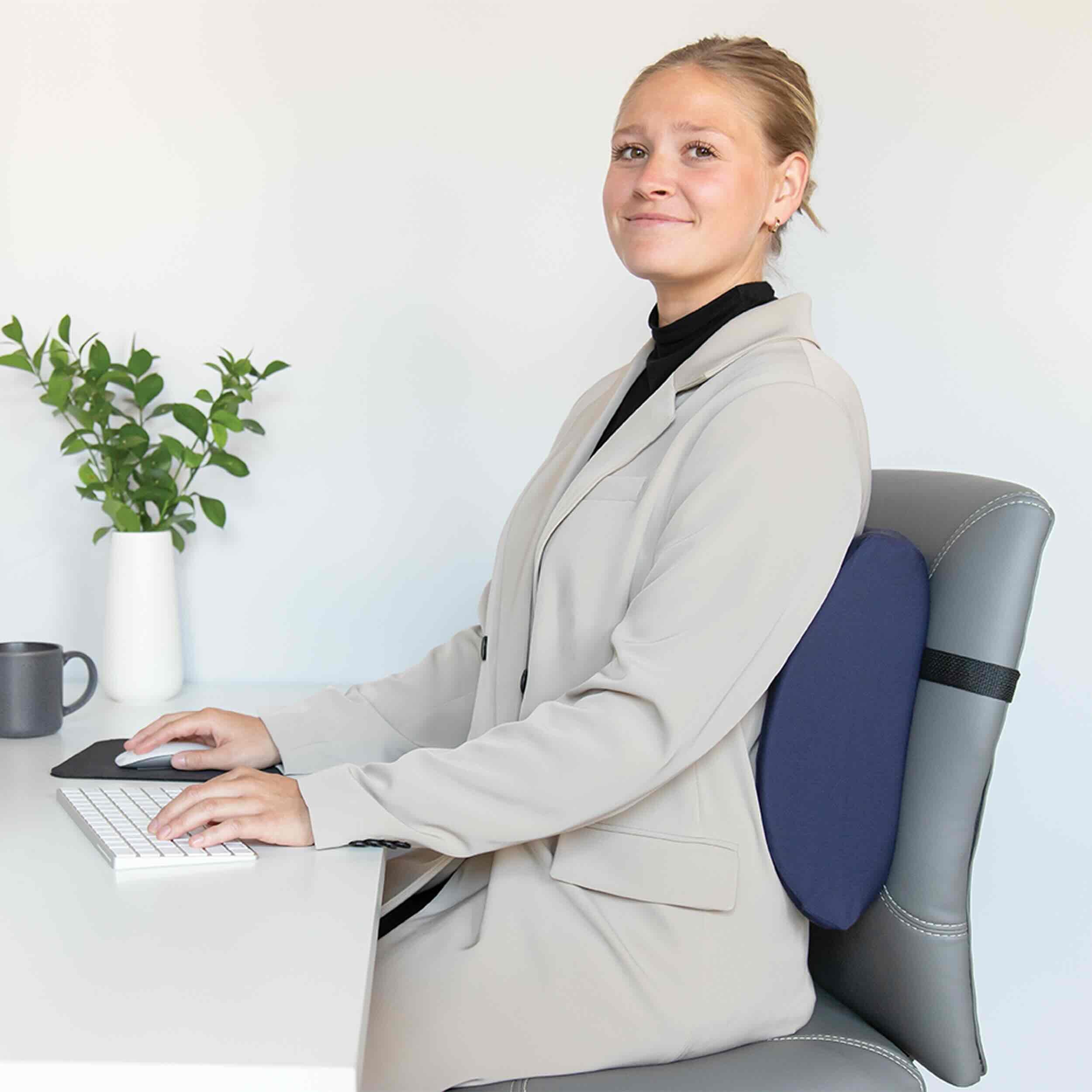 A woman sits at a desk using a keyboard and mouse, wearing a gray office chair with a navy blue lumbar support cushion behind her back. She is dressed in a light beige blazer and black turtleneck.