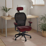 Ergonomic office chair in black mesh with burgundy leather seat, armrests, and headrest, positioned in a minimalist home office with a wood desk, lamp, framed art, and potted plants.