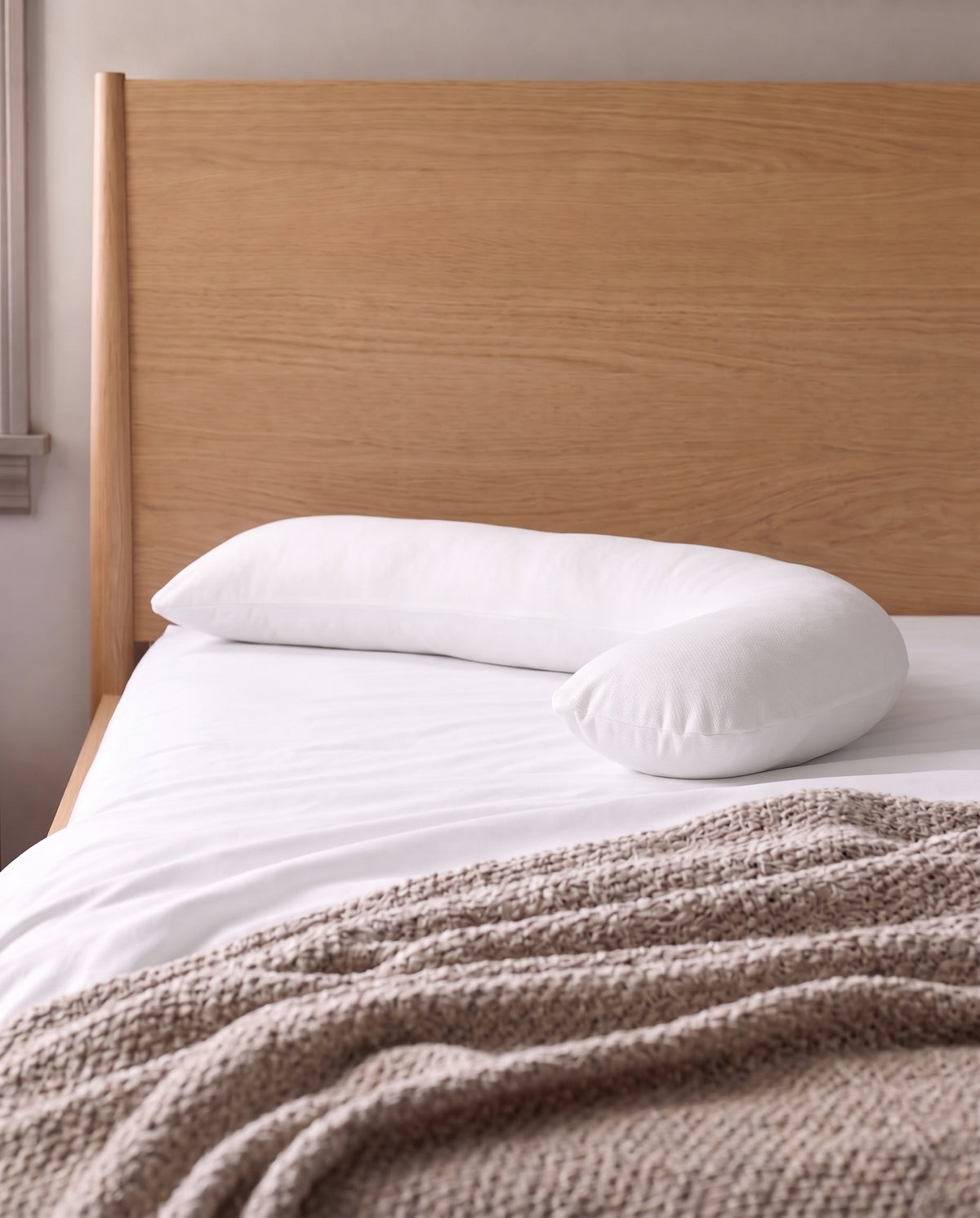 A white U-shaped pillow resting on a bed with white linens and a textured taupe throw blanket, set against a light wood headboard.