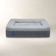 A square pet bed with a light gray plush top and a textured dark gray side, featuring a small blue 'Casper' brand tag on the front left side, set against a warm beige background.
