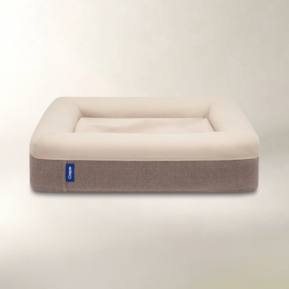 A rectangular pet bed with a cream-colored plush top and a taupe-brown fabric base, shown against a neutral light beige background. A small blue tag with the brand name 'Puppu' is visible on the left side.