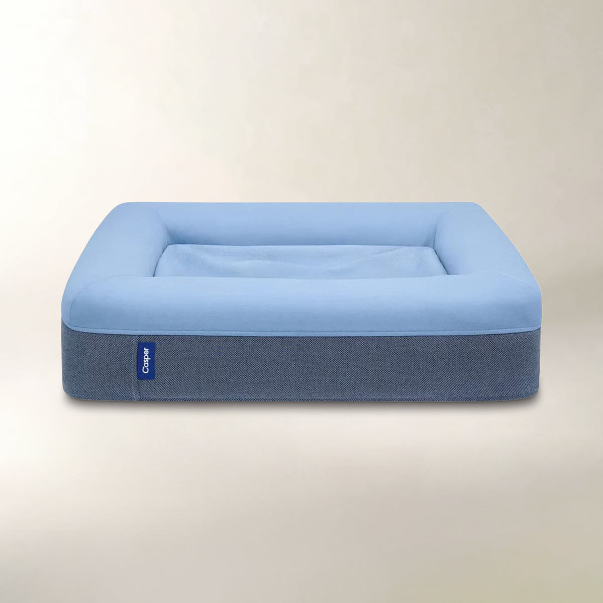 A square pet bed with a light blue top cushion and a textured dark blue-gray base, shown on a neutral beige background. A small blue tag on the left side of the base reads 'Cane'.