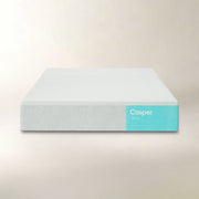 A Casper mattress shown from a front three-quarter angle against a warm beige background. The mattress has a light gray textured cover with a bright turquoise blue side label displaying the 'Casper' brand name.