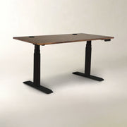 A modern standing desk with a rectangular brown wood-grain top and black metal T-style legs, shown on a neutral light gray studio background.