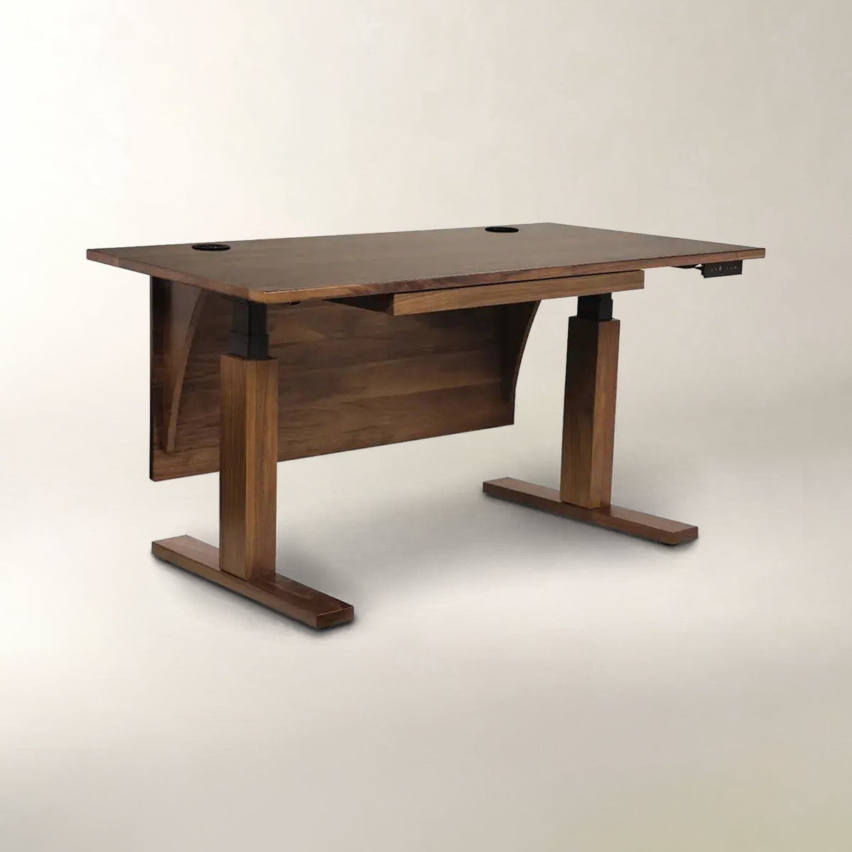 A modern wooden height-adjustable desk with a dark walnut finish, featuring a pull-out drawer, T-style legs, and cable management holes on the top surface, set against a neutral light gray background.