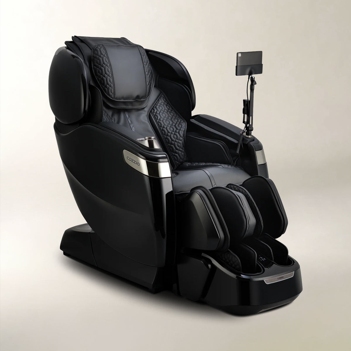 A black modern massage chair with a foot massager attachment and an adjustable armrest holding a tablet, photographed on a clean light beige studio background.
