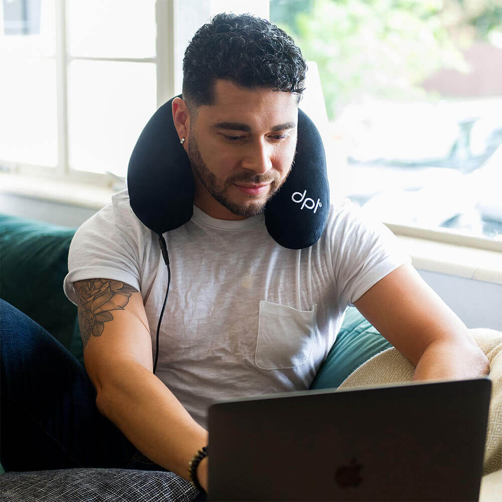 Man using a laptop with a neck pillow labeled 'dpt' on a couch.