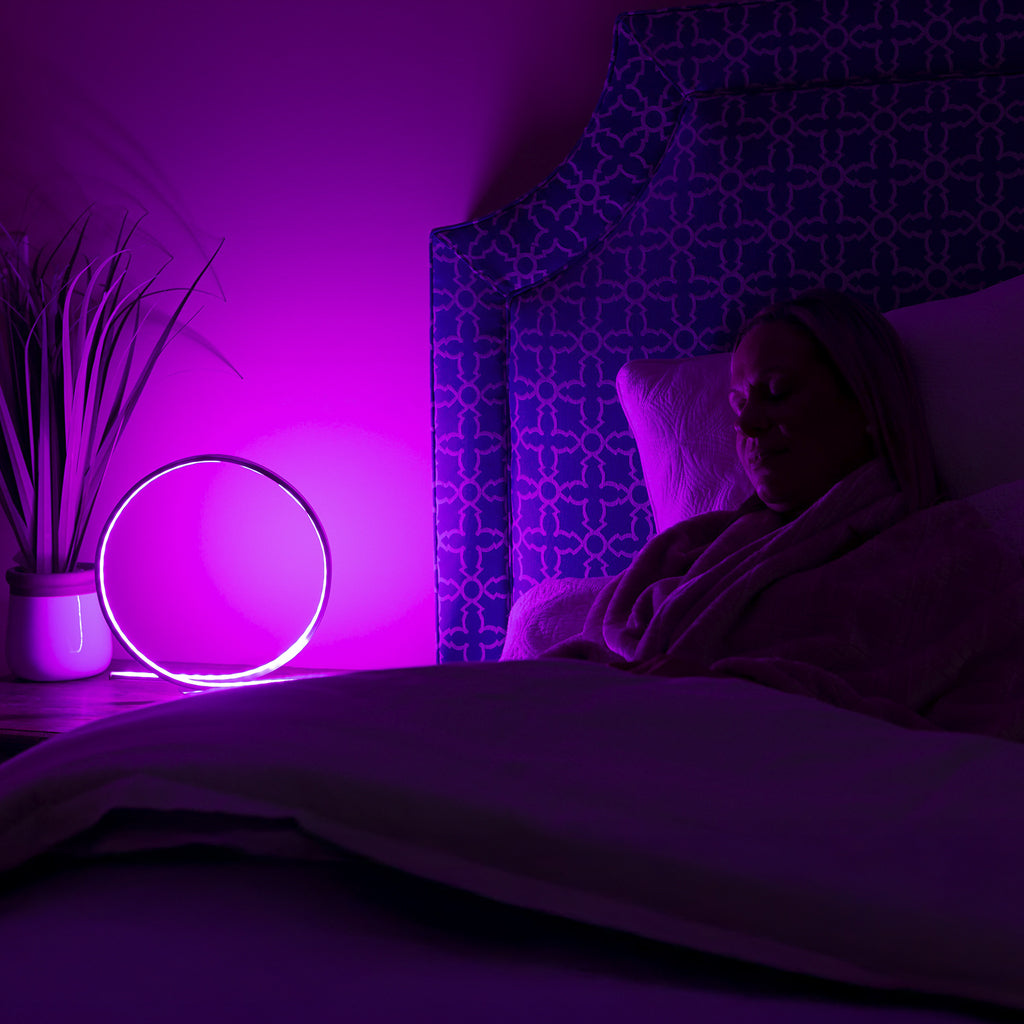 Person lying in bed under a blanket with a purple light illuminating the room.