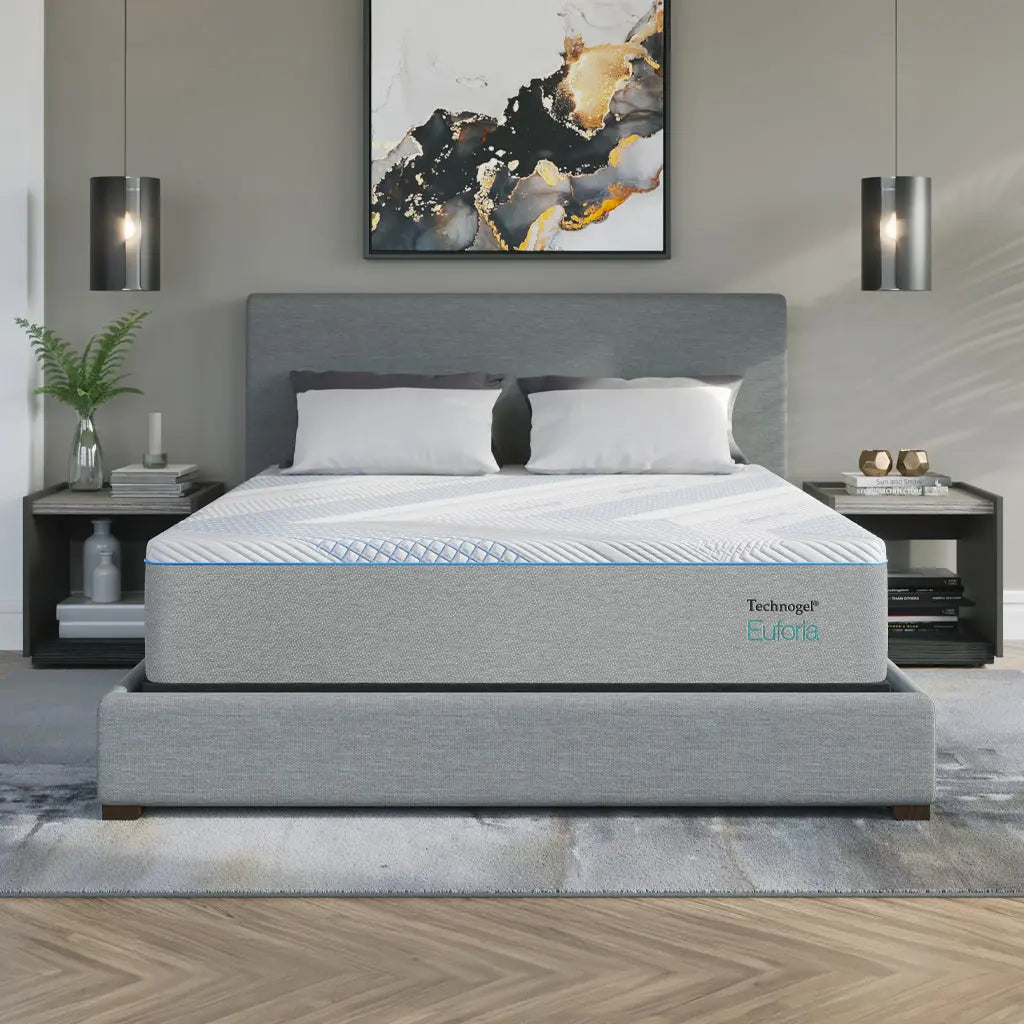 A Technogel® Euforia mattress sits atop a modern upholstered bed frame in a stylish bedroom. The mattress features a light gray side panel with the Technogel® Euforia logo and a white top cover with blue cooling accents. The room includes pendant lights, abstract artwork, and a soft neutral color palette.