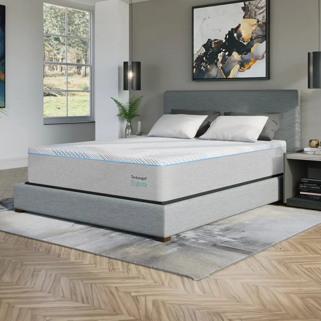 A modern bedroom features a Technogel® Euforia mattress on a low-profile, upholstered bed frame in light gray. The mattress has a sleek, breathable cover with blue accents and is topped with crisp white pillows. The space is styled with abstract art, pendant lighting, and natural light from a large window.