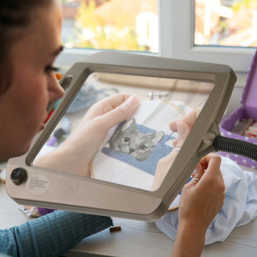 Person using a magnifying lamp to work on a craft project with a small cat illustration.