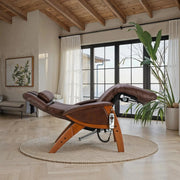 Brown leather zero gravity recliner with wood base positioned in a sunlit room featuring large windows, a round woven rug, and modern natural decor.