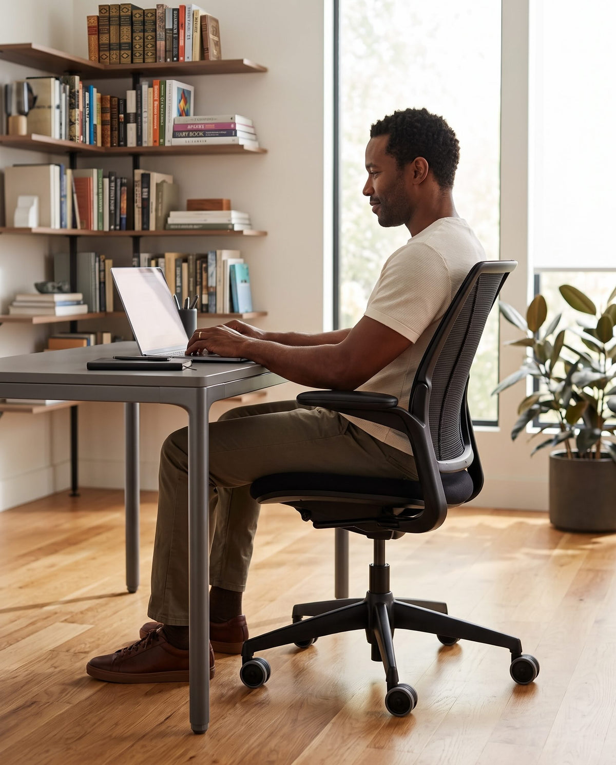 A man sits in a black mesh office chair at a gray desk, working on a laptop in a bright home office with bookshelves and a plant.