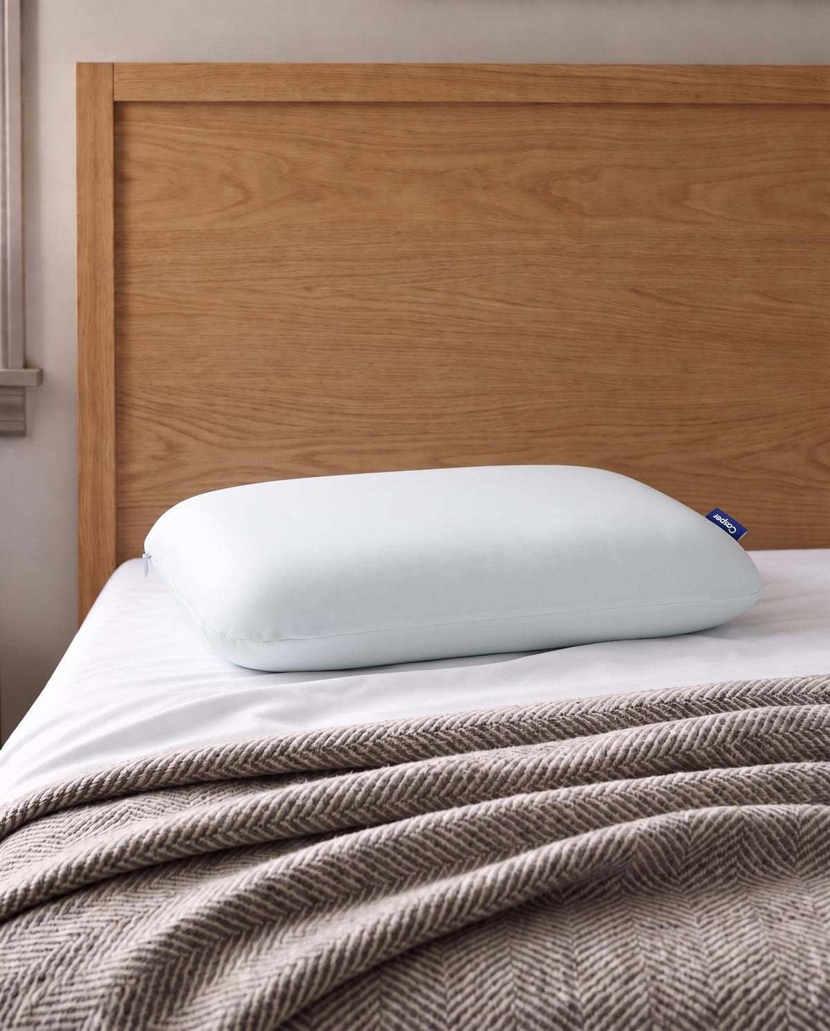 A light blue pillow resting on a bed with white sheets and a brown herringbone blanket, set against a light wood headboard.