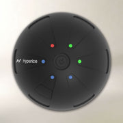 A top-down view of a black Hyperice massage ball on a light beige background. The textured ball features segmented panels, a central power button, and four colored LED indicator lights (red, green, blue) with the Hyperice brand logo on the left side.