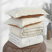 A stack of folded bedding, including two cream pillowcases with tan undersides, a cream quilt, and a white duvet, arranged on a round woven ottoman in a softly lit room.
