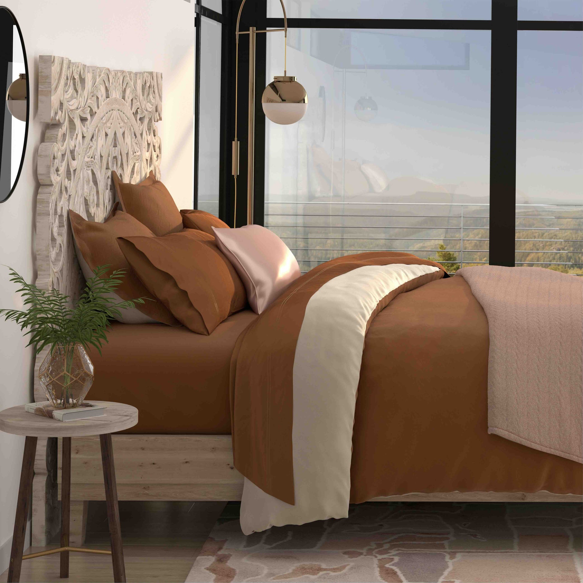 A styled bedroom scene showing a bed dressed in warm rust-brown bedding with a cream center panel and matching pillows, placed in a modern room with large windows, a side table, and wall decor.