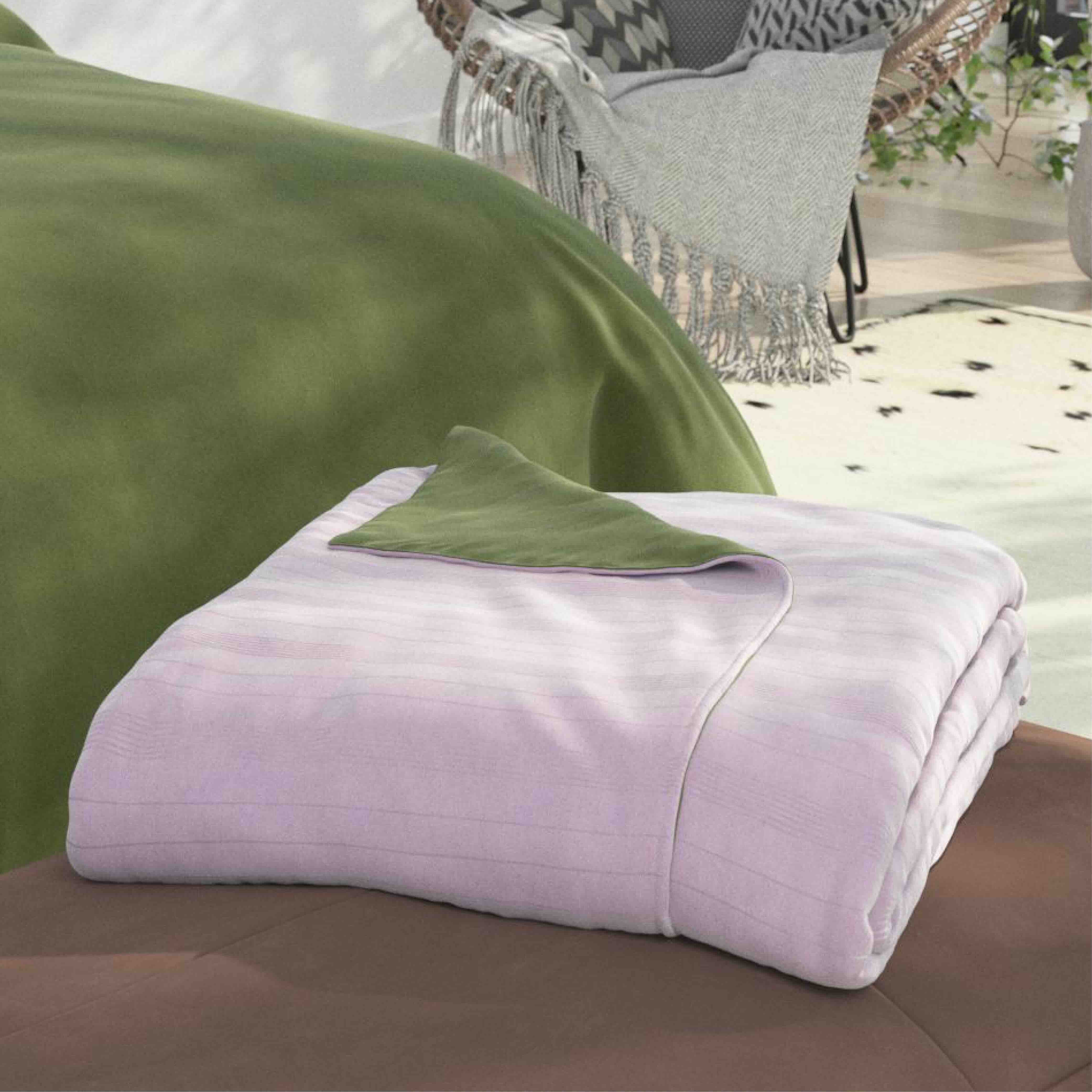 A folded light lavendar blanket with a matching green accent pillow is displayed on a brown sofa, with a green throw blanket draped over a chair in the background.