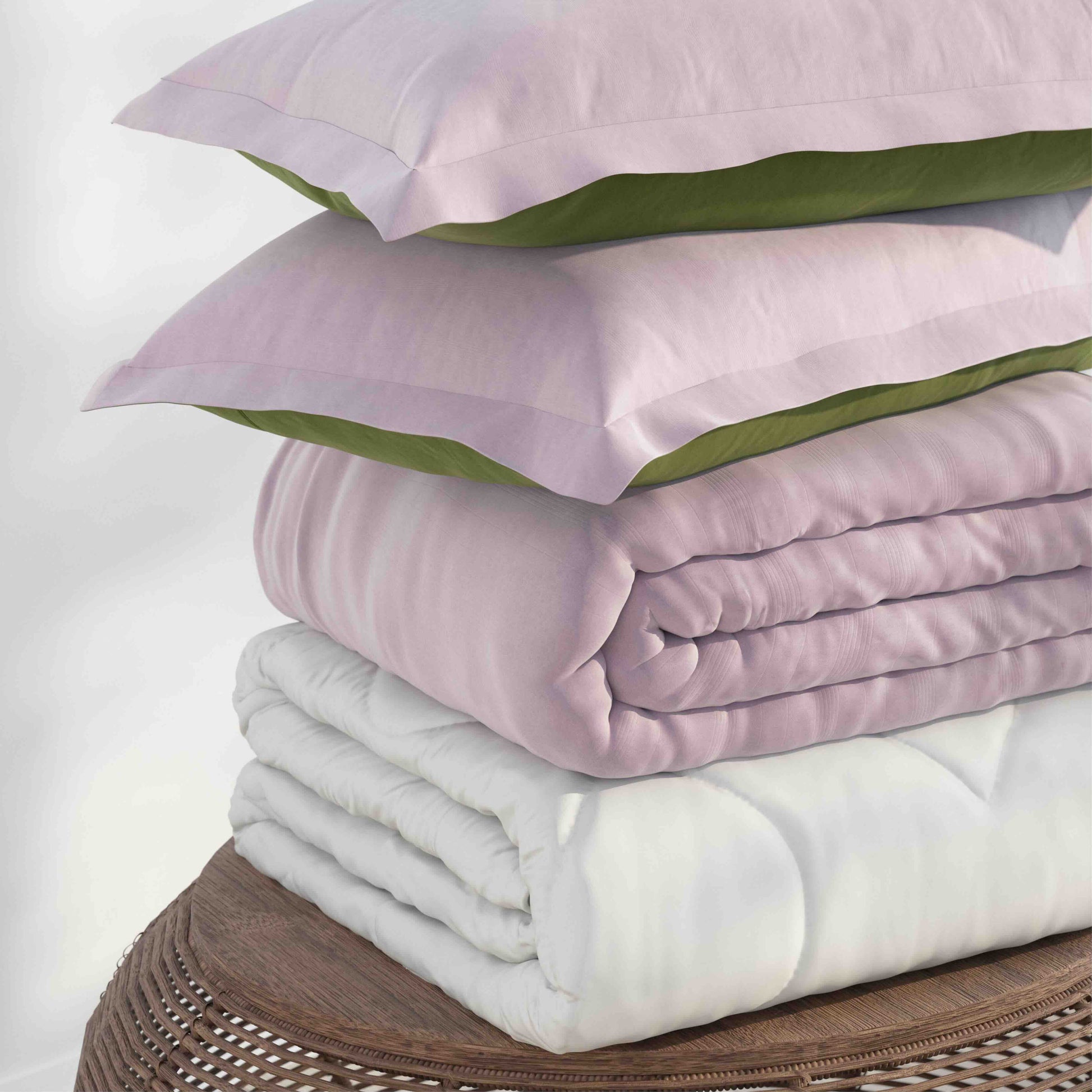 A stack of bedding including two light pink pillows with olive green undersides, a folded light pink quilt, and a folded white duvet, displayed on a round woven wooden stool against a white background.
